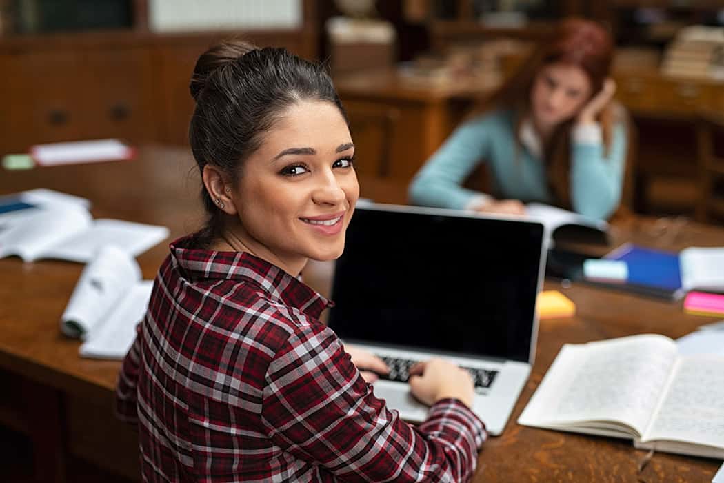 Female student sat at a desk in a library working on a laptop turns back to look at the camera and smiles